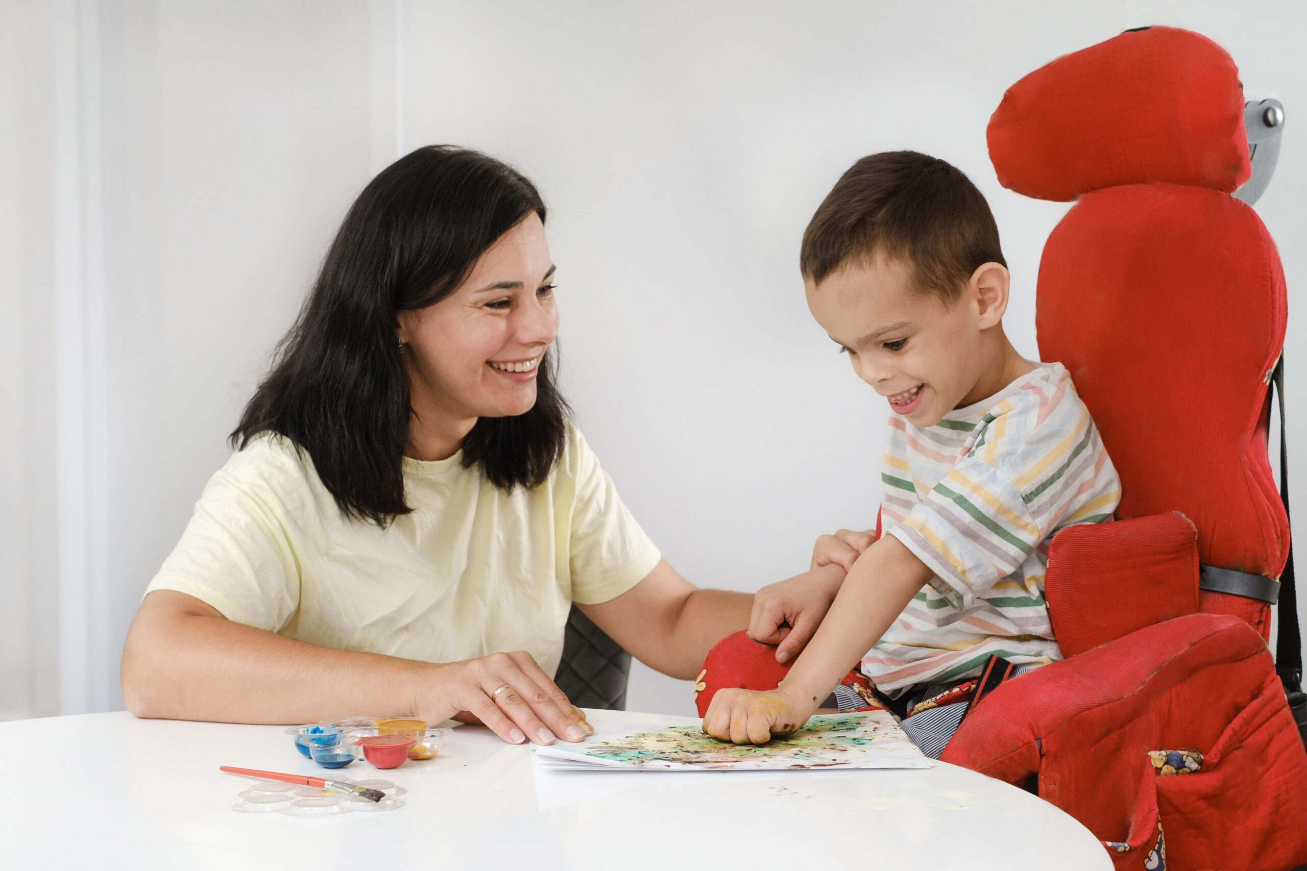 Young NDIS Child with cerebral palsy painting with fingers and hands. Happy handicap boy developing fine motor skills. Education for children with mental and physical disorders, sensory art therapy game